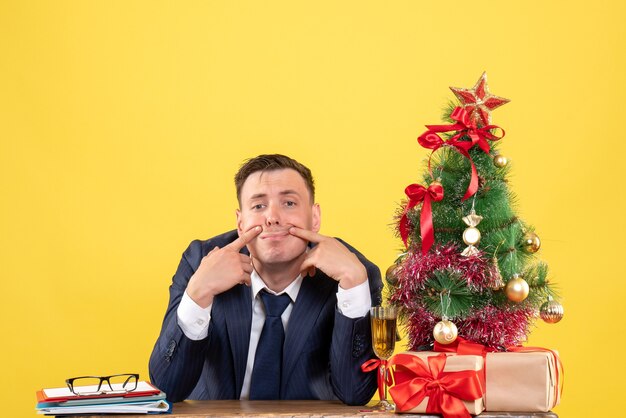 jovem tentando sorrir sentado à mesa perto da árvore de natal e presentes em amarelo