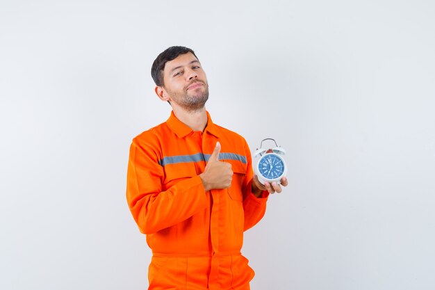 Jovem trabalhador aparecendo o polegar, segurando o despertador de uniforme e parecendo confiante.