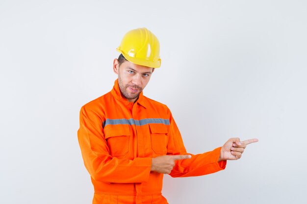 Jovem trabalhador apontando para o lado e sorrindo de uniforme, capacete.