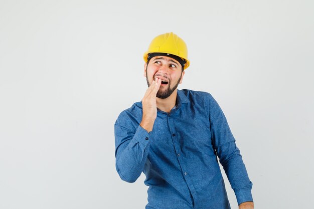 Jovem trabalhador com dor de dente dolorida na camisa, capacete e parecendo desconfortável