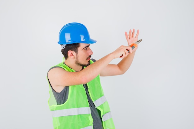 Foto grátis jovem trabalhador segurando um alicate e esticando a mão em direção a ele com uniforme de construção e olhando focado