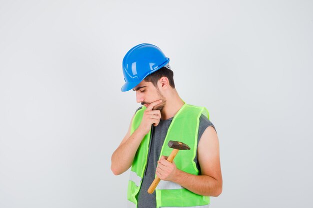 Jovem trabalhador segurando um machado em uma das mãos e parado em uma pose pensativa com uniforme de construção e olhando pensativo