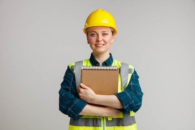 Jovem trabalhadora da construção civil sorridente usando capacete e colete de segurança abraçando o bloco de notas