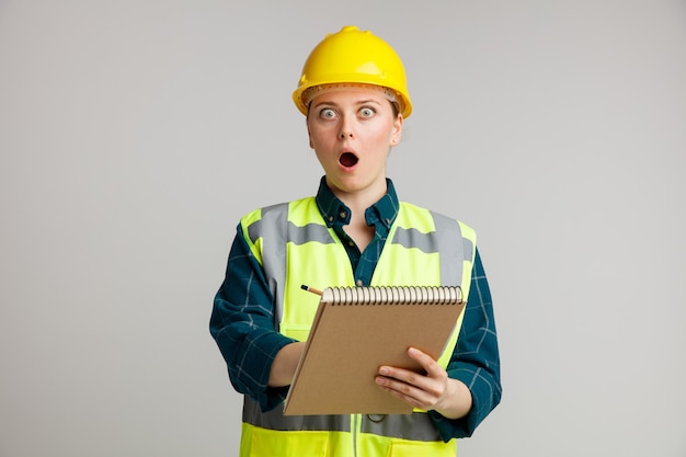 Jovem trabalhadora da construção civil surpreendida usando capacete e colete de segurança segurando um bloco de notas apontando um lápis sobre ele