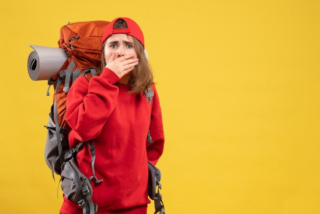 Jovem turista agitado de frente com mochila e boné vermelho em pé na parede amarela