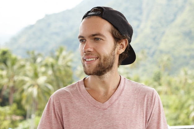 Jovem turista atraente e sorridente de boné preto virado para trás, aproveitando o clima ensolarado e os dias quentes de verão durante as férias em um país tropical