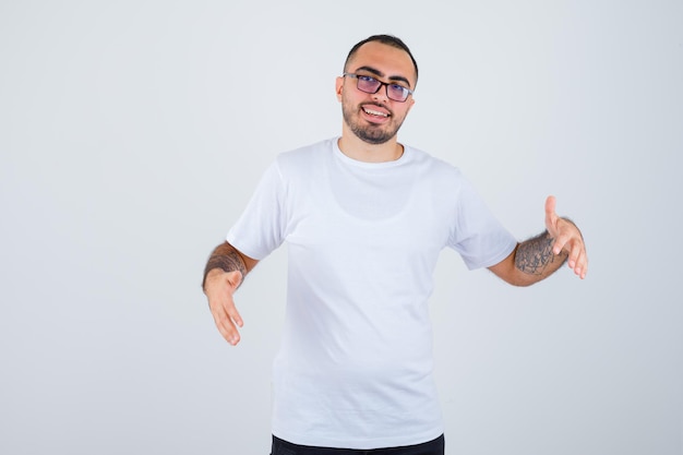 Jovem usando óculos e esticando as mãos segurando algo em uma camiseta branca e calça preta e parecendo feliz