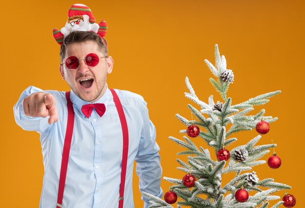 Jovem usando suspensórios gravata borboleta na borda com óculos de Papai Noel e vermelho, em pé ao lado da árvore de Natal, rindo e apontando com o dedo indicador para você sobre a parede laranja
