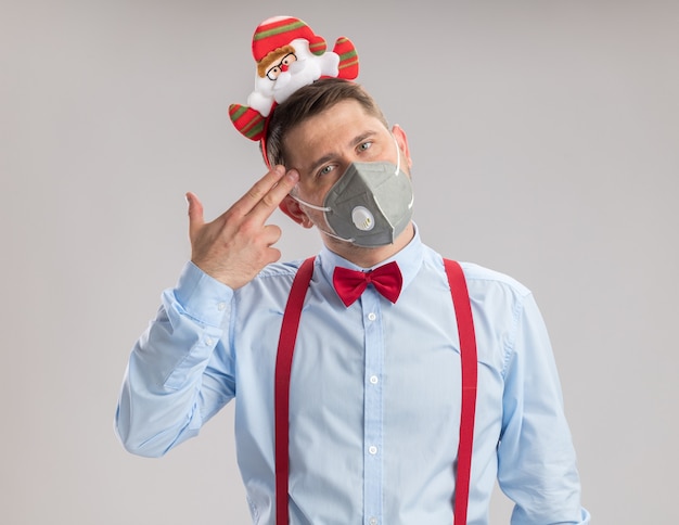 Jovem usando suspensórios gravata-borboleta na borda com papai noel usando máscara facial protetora olhando para a câmera fazendo gesto de pistola com os dedos sobre o templo em pé sobre fundo branco