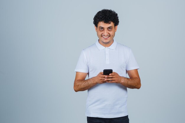 Jovem usando telefone celular em t-shirt branca e olhando alegre, vista frontal.