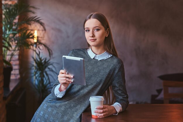 Jovem vestida com um elegante vestido cinza segura uma xícara de café para viagem e segura um tablet enquanto se inclina sobre a mesa em uma sala com interior loft, olhando para uma câmera.