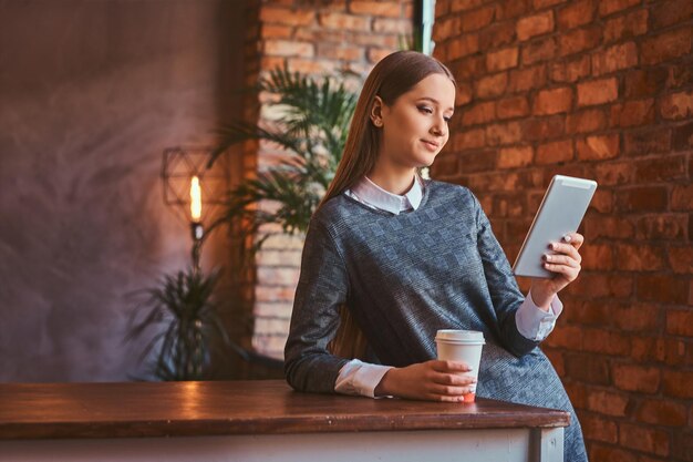 Jovem vestida com um elegante vestido cinza segura uma xícara de café para viagem e usando um tablet enquanto se apoia na mesa em uma sala com interior loft.