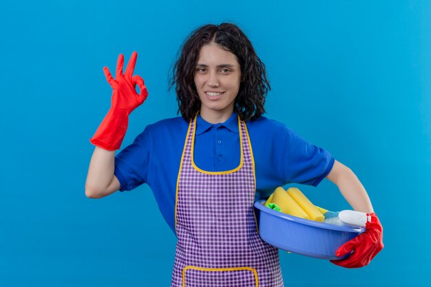 Jovem vestindo avental e luvas de borracha, segurando a bacia com ferramentas de limpeza sorrindo amigável fazendo ok assinar sobre parede azul