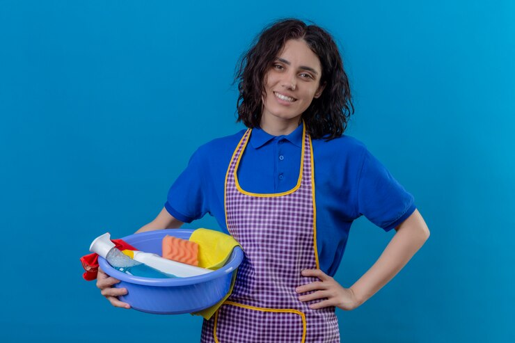 Foto grátis jovem vestindo avental segurando a bacia com ferramentas de limpeza olhando confiante sorridente alegre parede azul