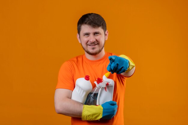 Jovem vestindo camiseta laranja e luvas de borracha segurando material de limpeza sorrindo positivamente