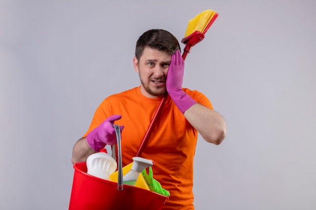 Jovem vestindo camiseta laranja e luvas de borracha segurando um balde com ferramentas de limpeza e o esfregão confuso tocando a cabeça por engano em pé sobre uma parede verde