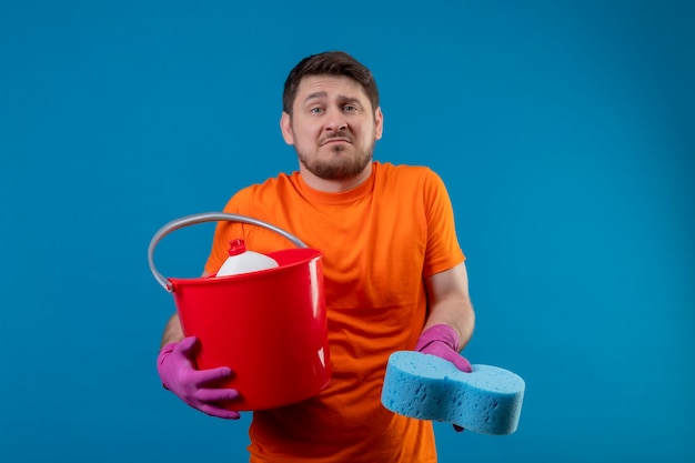 Foto grátis jovem vestindo camiseta laranja e luvas de borracha segurando um balde com ferramentas de limpeza e uma esponja