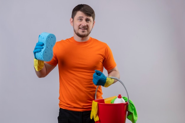 Jovem vestindo camiseta laranja e luvas de borracha segurando um balde com ferramentas de limpeza e uma esponja