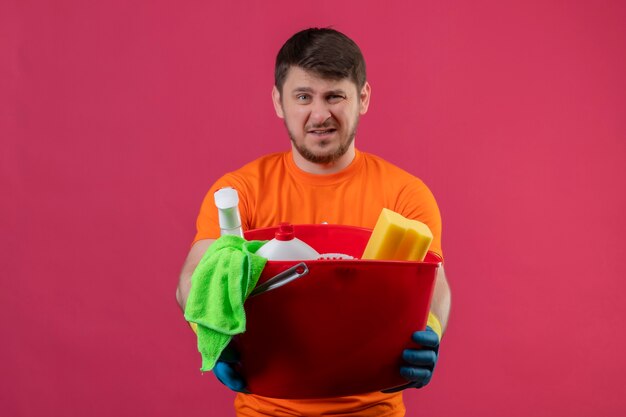 Jovem vestindo camiseta laranja e luvas de borracha segurando um balde com ferramentas de limpeza