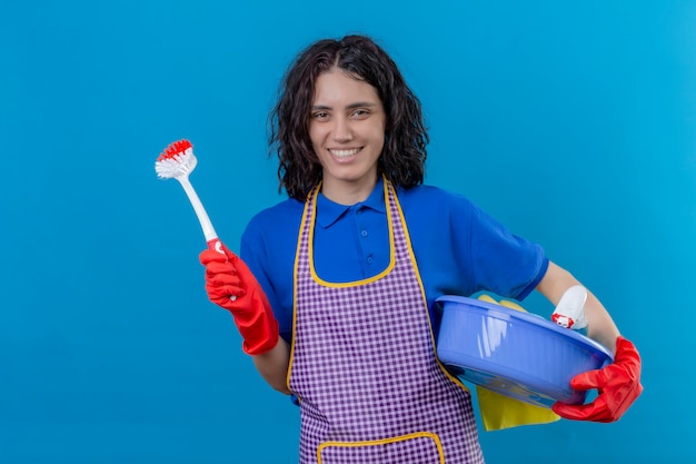 Jovem vestindo luvas de borracha e avental segurando a bacia com ferramentas de limpeza e escova com grande sorriso no rosto, positivo e feliz sobre parede azul