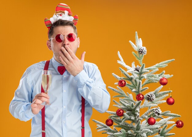 Jovem vestindo suspensórios gravata borboleta na borda com óculos de papai noel e óculos vermelhos ao lado da árvore de natal segurando uma taça de champanhe