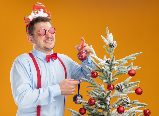 Foto grátis jovem vestindo suspensórios gravata borboleta na borda com óculos de papai noel e vermelhos, ao lado da árvore de natal, parecendo confuso segurando brinquedos para a árvore sobre fundo laranja
