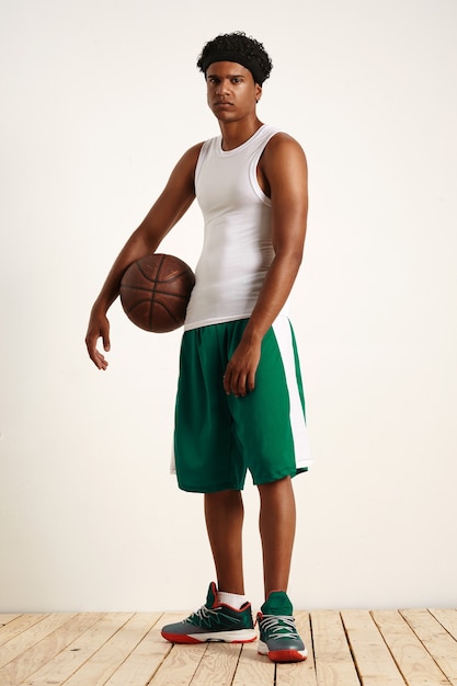 Foto grátis jovem vestindo uma camisa branca, bandana, shorts verdes e tênis verdes em pé com uma velha bola de basquete de couro debaixo do braço