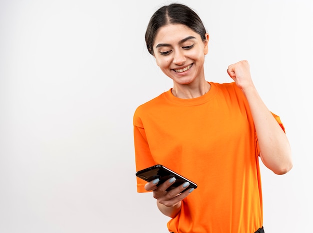 Jovem vestindo uma camiseta laranja segurando um smartphone com o punho cerrado, feliz e animada, sorrindo alegremente e regozijando-se por seu sucesso de pé sobre uma parede branca