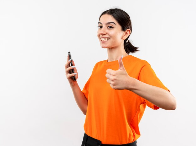 Jovem vestindo uma camiseta laranja segurando um smartphone olhando para o lado e sorrindo alegremente mostrando os polegares em pé sobre uma parede branca