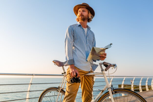 Jovem viajando de bicicleta à beira-mar nas férias de verão à beira-mar no pôr do sol, segurando um mapa turístico com a câmera