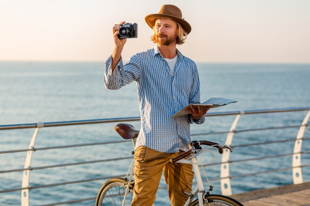 Jovem viajando de bicicleta à beira-mar nas férias de verão à beira-mar no pôr do sol, segurando um mapa turístico, tirando foto na câmera