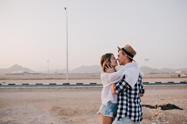 Jovem viajante com chapéu elegante e mulher com roupa da moda bonita beijando perto da estrada no fim de semana. jovem adorável abraçando suavemente seu lindo namorado e sorrindo para a natureza