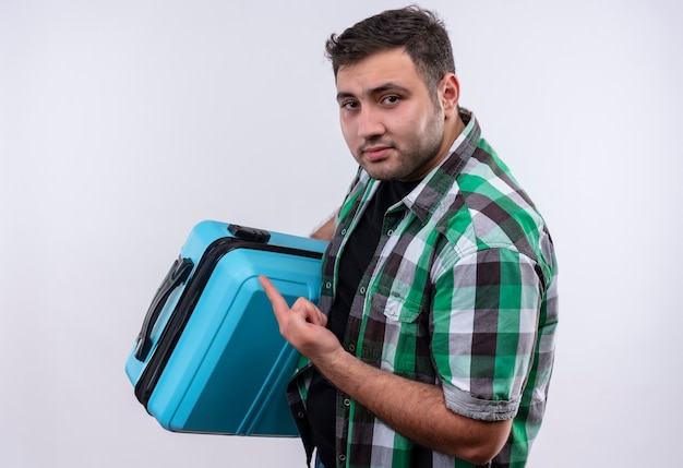 Jovem viajante de camisa xadrez segurando uma mala apontando com o dedo e parecendo confiante em pé sobre uma parede branca