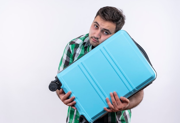 Jovem viajante de camisa xadrez segurando uma mala com uma expressão triste no rosto em pé sobre uma parede branca