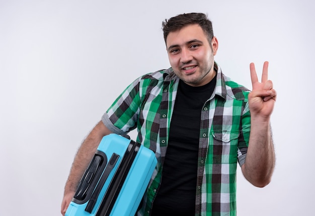 Jovem viajante de camisa xadrez segurando uma mala, sorrindo e mostrando o número três com os dedos em pé sobre uma parede branca