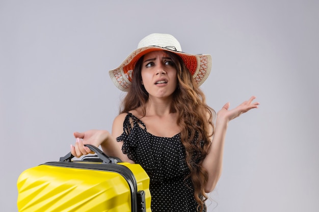 Jovem viajante linda em um vestido de bolinhas com chapéu de verão segurando uma mala sem noção e confusa, sem resposta, em pé com os braços levantados sobre um fundo branco
