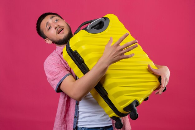 Jovem viajante segurando uma mala amarela parecendo confuso em pé sobre um fundo rosa