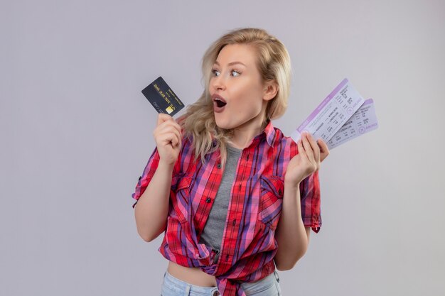Jovem viajante surpresa, vestindo camisa vermelha, segurando um cartão de crédito e bilhetes na parede branca isolada