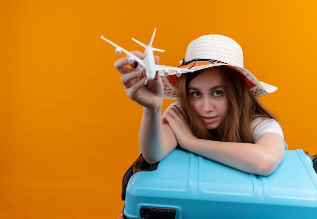 Jovem viajante usando um chapéu esticando o modelo do avião e colocando o braço na mala na parede laranja isolada com espaço de cópia