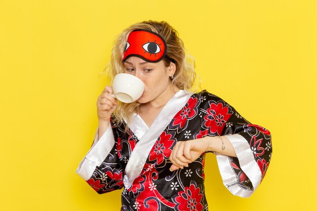 Jovem, vista frontal, mulher com roupão de noite e máscara de olho se preparando para dormir, bebendo café na cama de noite amarela de modelo feminino de sono