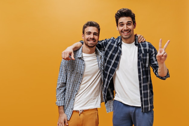 Jovens alegres em camisas xadrez azuis, camisetas brancas e calças coloridas posam na parede laranja de ótimo humor e sorriem.