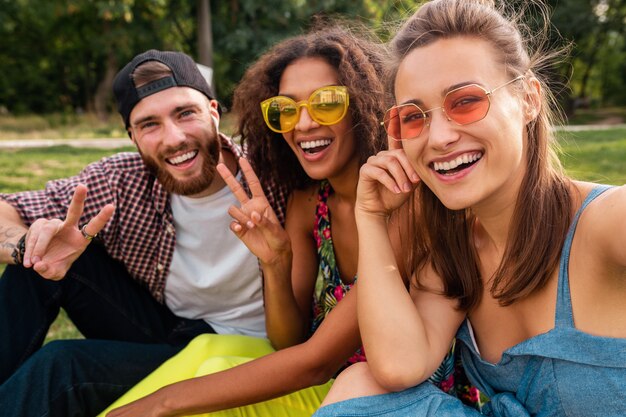 Jovens amigos elegantes e felizes sentados no parque, fazendo selfie