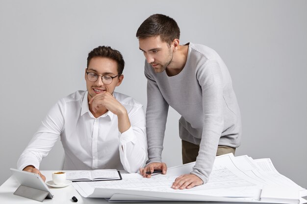 Jovens arquitetos elegantes trabalhando juntos em um novo projeto habitacional, verificando desenhos e usando tablet digital