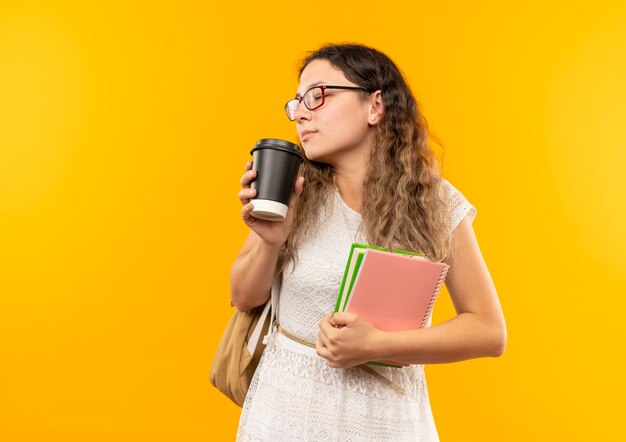 Jovens bonitas colegiais de óculos e mochila segurando um livro e um bloco de notas e cheirando a xícara de café com os olhos fechados, isolada em um fundo amarelo com espaço de cópia