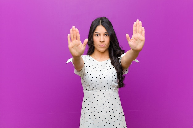 Jovens bonitas mulher latina olhando sério com as duas palmas das mãos abertas contra a parede roxa