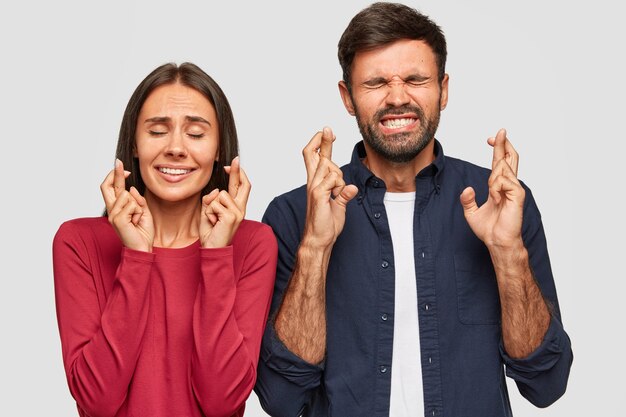 Jovens do sexo masculino e feminino fecham os olhos, cruzam os dedos de esperança, antecipam algo com expressões fiéis, têm sorrisos