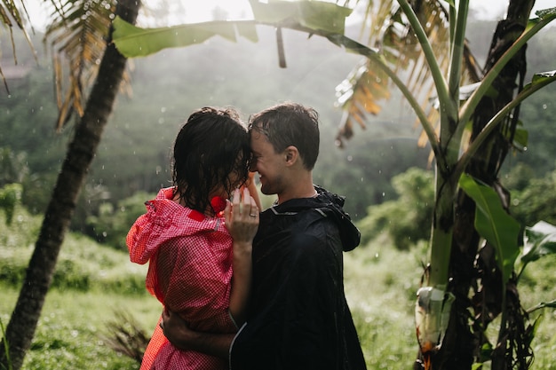 Jovens em capas de chuva, abraçando a natureza. Foto ao ar livre de casal romântico beijando na floresta tropical.