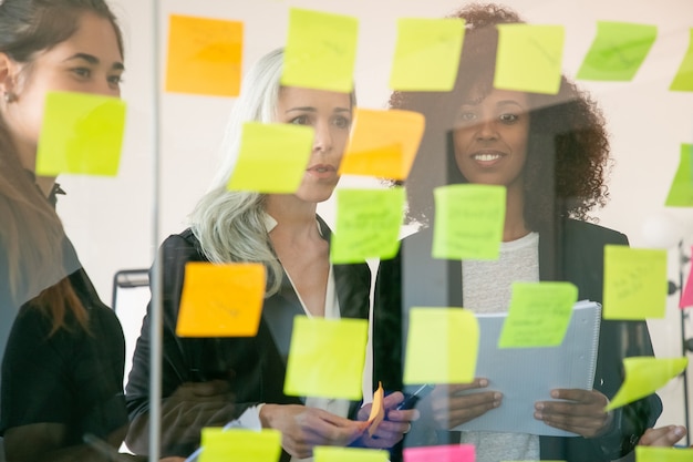 Jovens empresários contentes, discutindo o plano de marketing e fazendo anotações em adesivos. Colegas bem-sucedidos e confiantes de terno reunidos na sala do escritório. Conceito de trabalho em equipe, negócios e brainstorm