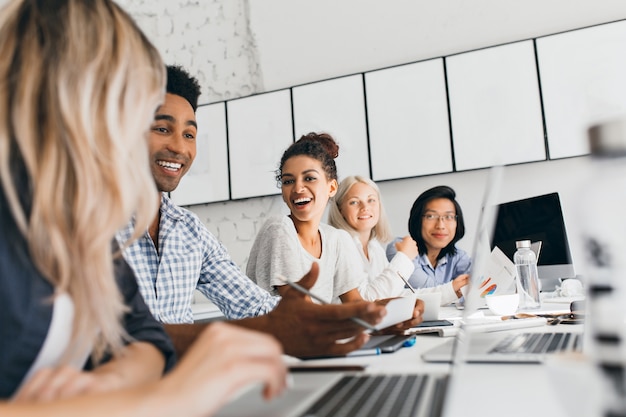 Jovens empresários discutindo algo com um sorriso durante a conferência. Retrato interno de funcionários internacionais sentados no escritório com laptops e falando sobre trabalho.