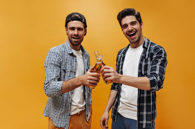 Foto grátis jovens morenos felizes e emocionais em camisas xadrez azuis sorriem e brindam com garrafas de cerveja em fundo laranja isolado